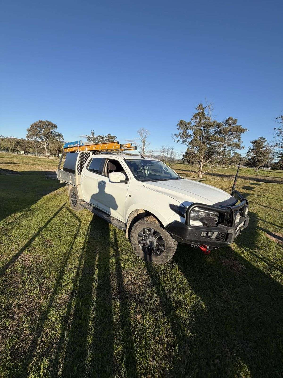Kayden's white dual-cab ute with ladders on the rack, parked in a paddock with gum trees