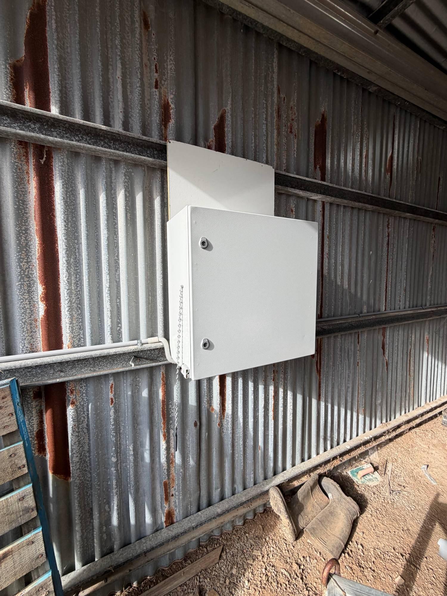 New white electrical enclosure mounted on a backing board on a corrugated iron shed wall, with conduit run in from below
