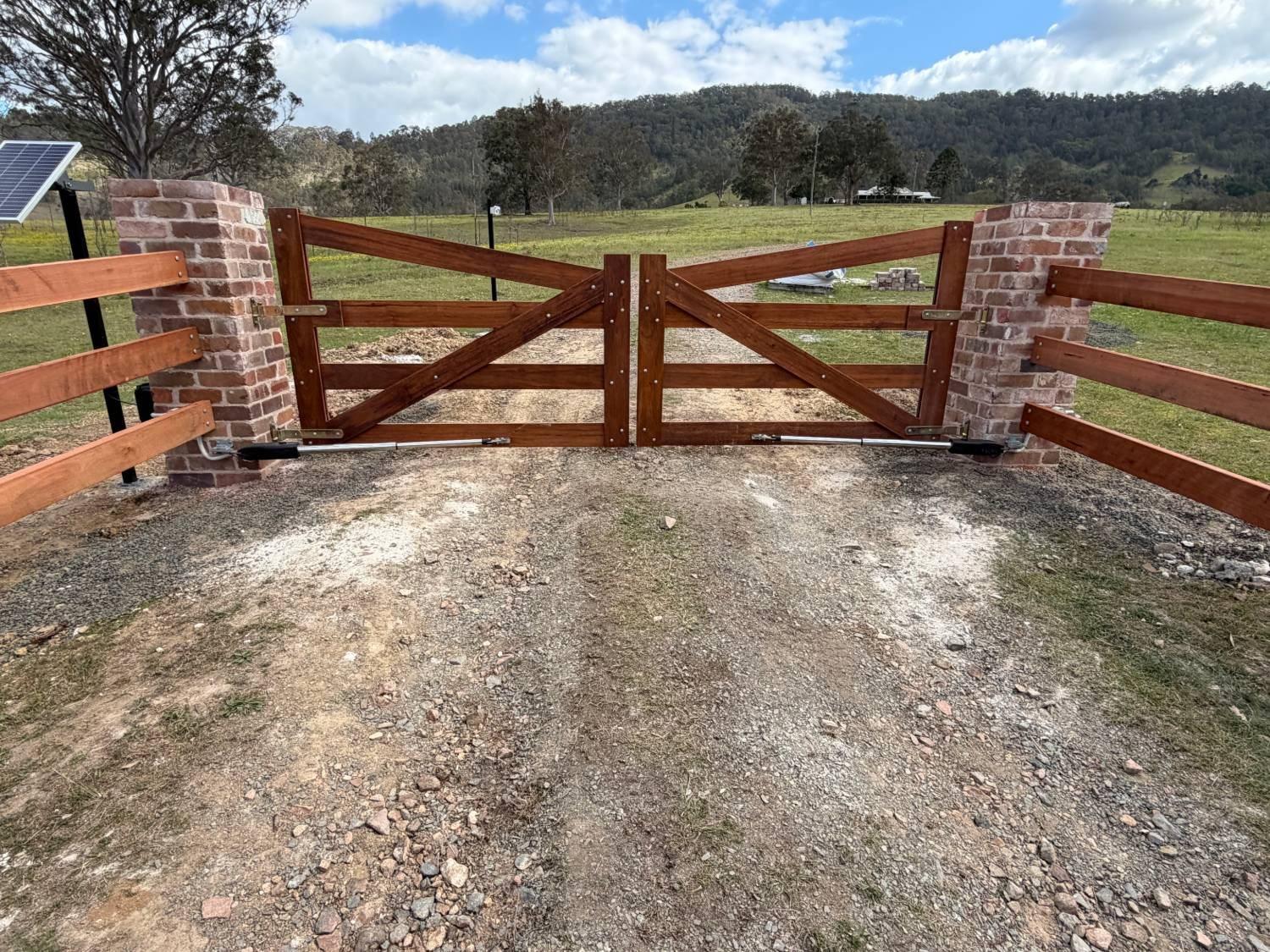 Timber farm gates between brick pillars at a property entrance, with automatic gate openers fitted and hills behind