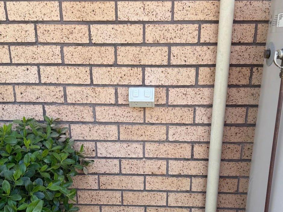 Weatherproof double power point mounted on a brick wall beside a downpipe and hot water system