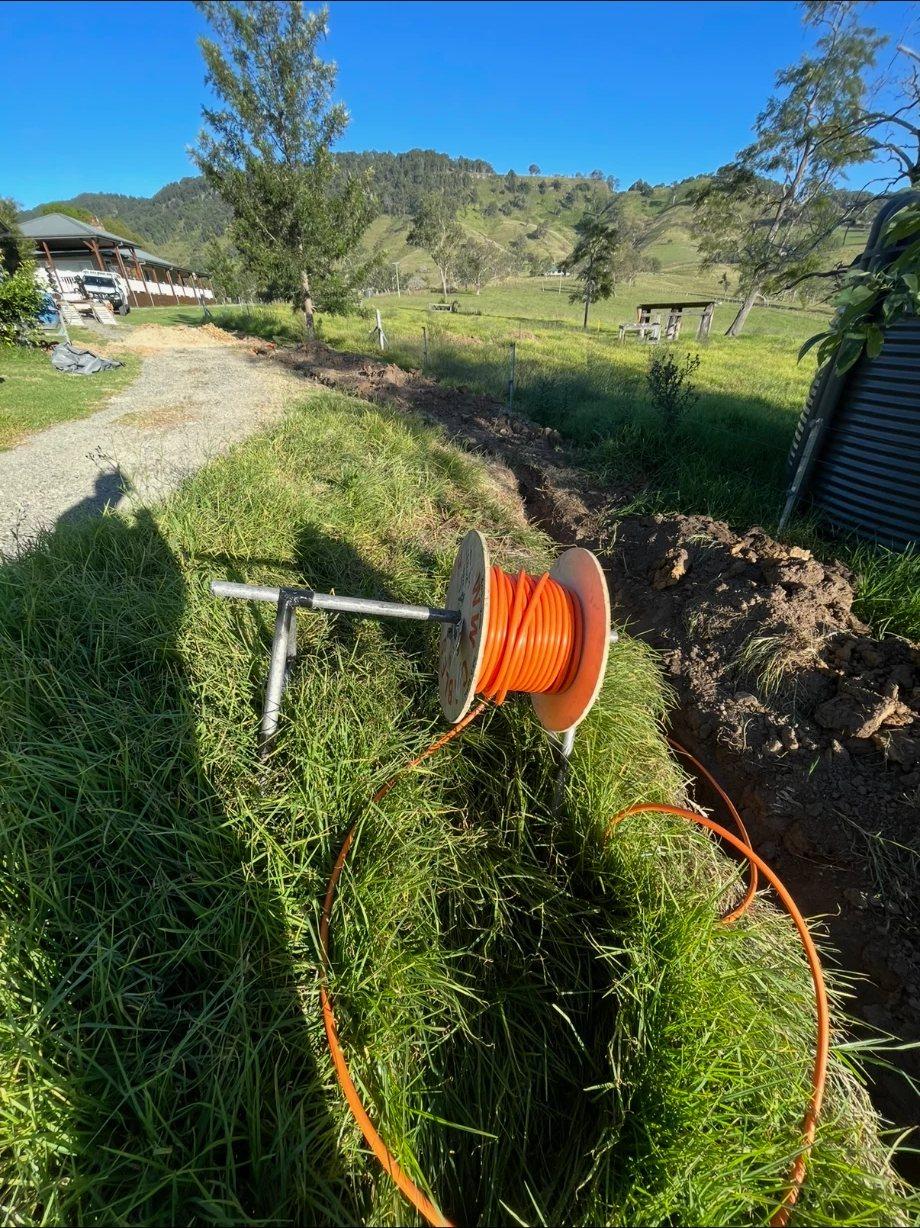 Orange conduit coming off a reel into an open trench beside a farm driveway, with hills behind