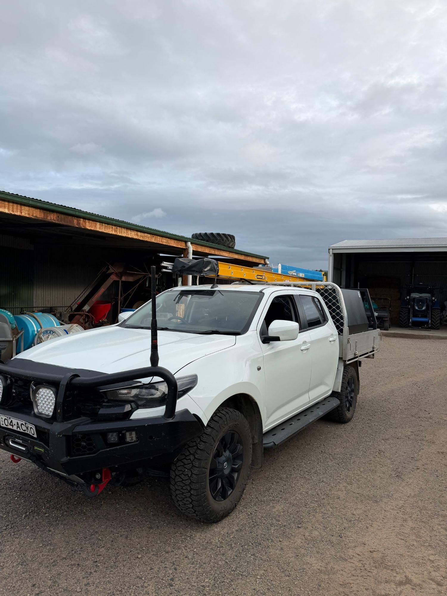 Kayden's white dual-cab ute with ladders on the rack, parked between farm sheds with tractors and machinery behind