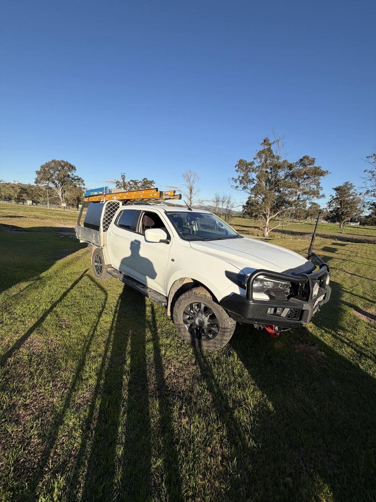 Kayden's white dual-cab ute with ladders on the rack, parked in a paddock with gum trees
